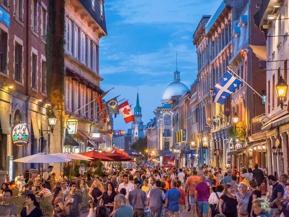 Rue piétonne bondée du Vieux-Montréal à la tombée du jour, terrasses, drapeau du Québec et dôme argenté au fond