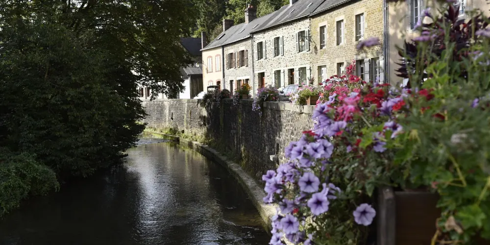 Maisons de pierre alignées au bord de l’Ernée à Chailland, pétunias violets et roses fleurissant le quai en été